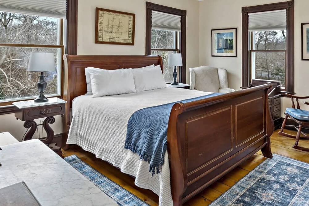 A cozy bedroom featuring a wooden sleigh bed, two lamps, and a comfortable armchair beside large windows.