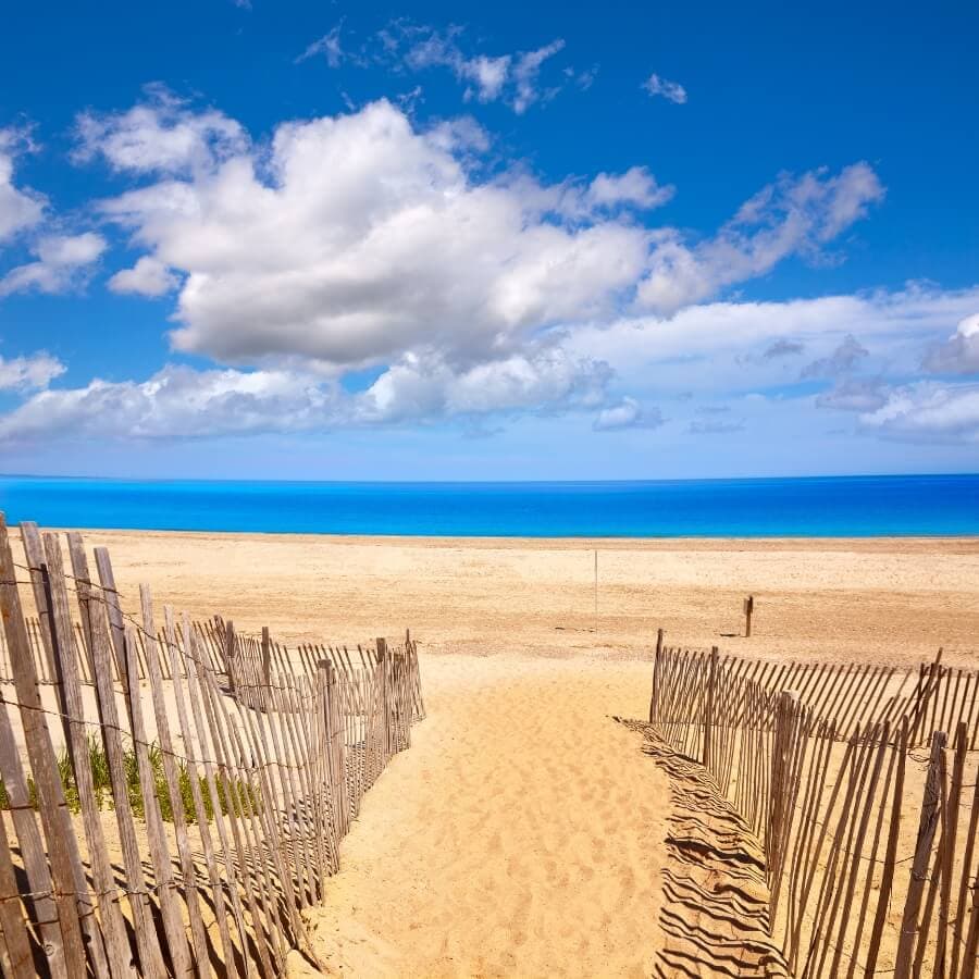 A sandy path leads to a tranquil beach under a bright blue sky with fluffy clouds.