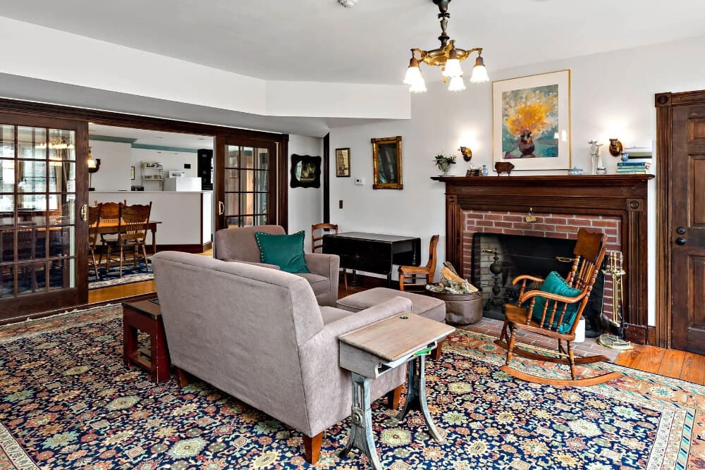 A cozy living room featuring a sofa, rocking chair, and a fireplace, with wooden accents and a colorful rug.